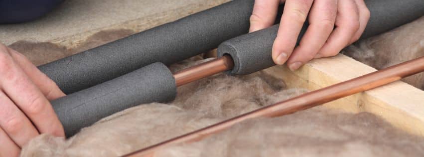 Hands applying foam pipe insulation to copper pipes surrounded by insulation material to prevent freezing.