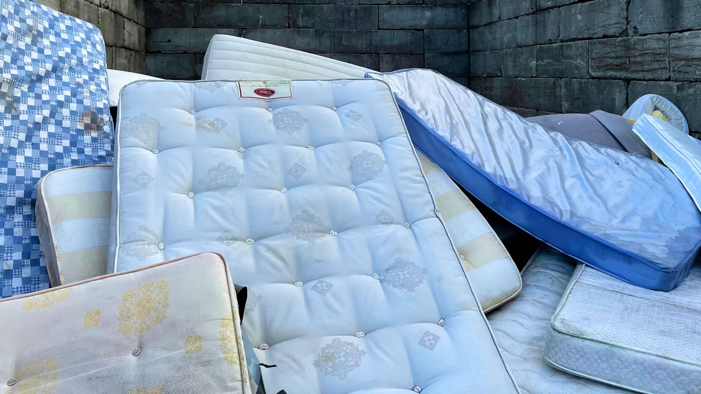 Pile of old mattresses stacked outdoors against a concrete wall, ready for disposal or recycling.